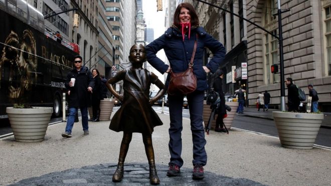 Женщина позирует рядом со статуей девушки, стоящей перед Уолл-Булл, в рамках кампании, проводимой американским управляющим фондом Стейт-стрит, чтобы подтолкнуть компании поставить женщин на свои доски в финансовом районе Нью-Йорка