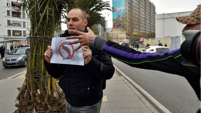 صورة لرجل يحمل لافتة للتعبير عن رفضة للـ"عهدة الخامسة" وشرطي في زي مدني يحاول انتزاعها