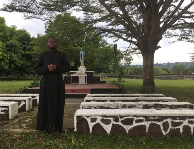 Отец Иосиф Мусубао носит черную рясу и стоит возле алтаря на открытом воздухе и скамей