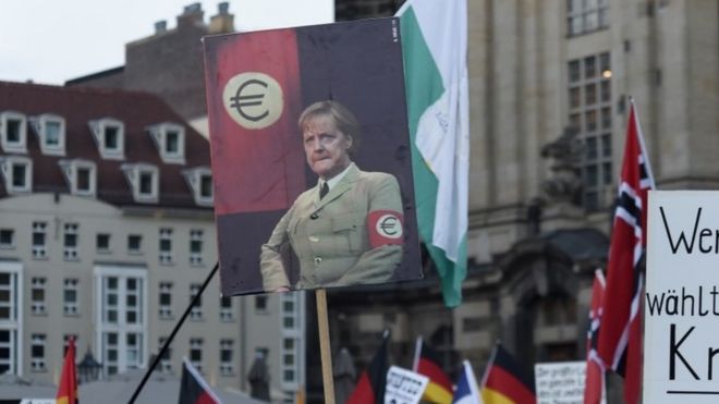 Протестующие держат плакат с изображением канцлера Германии Ангелы Меркель в форме нацистского стиля с эмблемой евро в качестве повязки на руке во время акции протеста в Пегиде (05 октября 2015 г.)