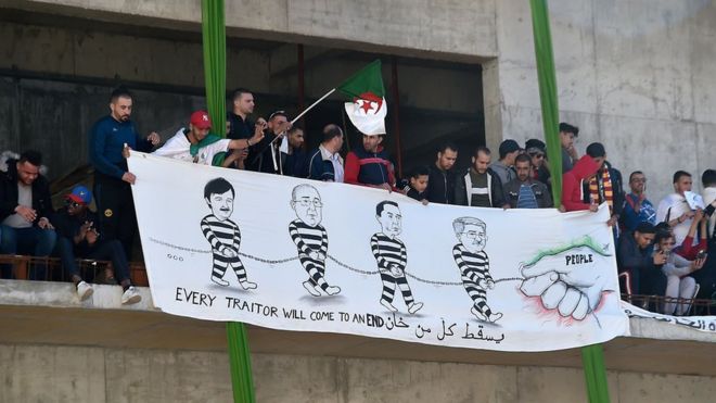 Люди держат плакат с призывом к президенту, генеральному секретарю Всеобщего профсоюза алжирских рабочих (UGTA), председателю Алжирского форума шеф-поваров (FCE) и бывшему премьер-министру «предатели».