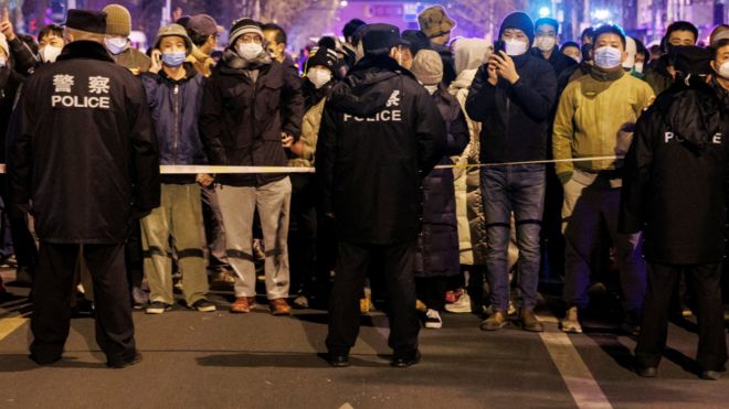 People stand behind a police cordon as they watch a protest over coronavirus disease (COVID-19) restrictions, following a commemoration of the victims of a fire in Urumqi, as outbreaks of COVID-19 continue, in Beijing, China, November 27, 2022.