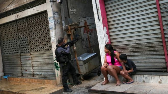 Sentados em frente a loja fechada, mãe e filho observam policial ao lado apontando arma grande