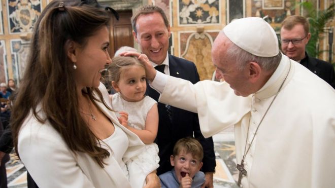 В роскошной обстановке папа Франциск, справа от рамки, кладет руку на голову молодой девушки, которую держат на руках улыбающаяся мать. Молодой мальчик - возможно, другой ребенок пары - смотрит снизу, его большой палец во рту.