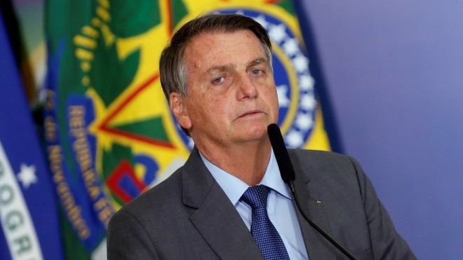 Bolsonaro com expressão séria falando ao microfone, com bandeiras no fundo