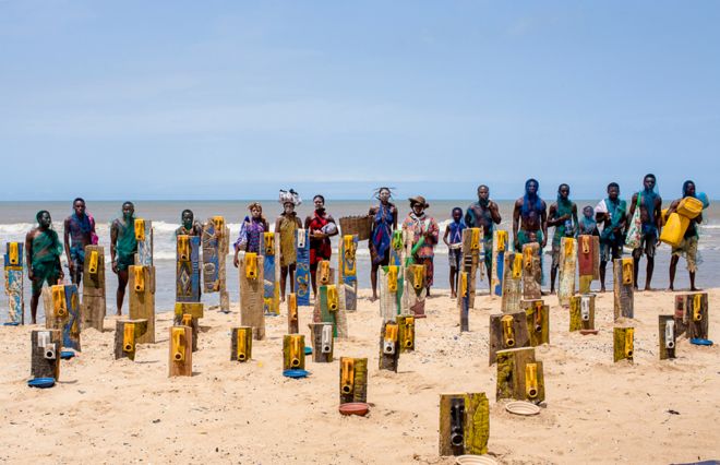 Люди стоят возле художественных работ с использованием канистр художника Сержа Аттуквея Клотти на пляже в Аккре, Гана