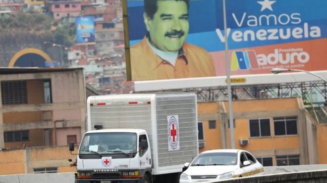 Рекламный щит с изображением президента Венесуэлы Николаса Мадуро виден на заднем плане, как грузовики, помеченные логотипом Международной федерации обществ Красного Креста и Красного Полумесяца (МФКК) и считающиеся перевозящими гуманитарную помощь, едут по шоссе в Каракасе, Венесуэла, апрель 16, 2019