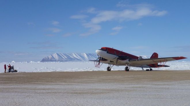 Красный самолет стоит на взлетно-посадочной полосе. Поле льда и горы видимые на заднем плане.