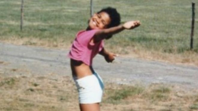Sara Jayne-King criança, sorrindo para foto e abrindo braços em ambiente ao ar livre