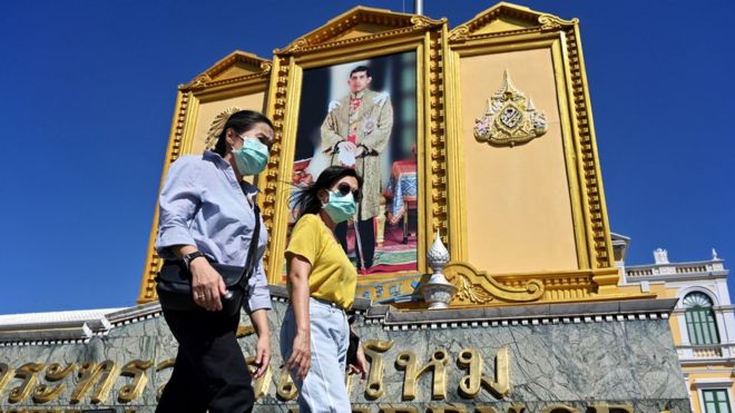 Люди в масках проходят мимо большого портрета короля Таиланда Маха Ваджиралонгкорна возле Большого дворца в Бангкоке 27 января 2020 года