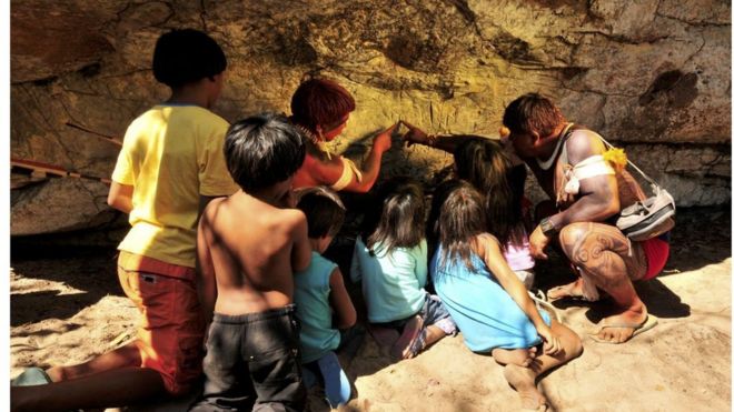 Registro de Índio waurá ensinando a mitologia em torno do guerreiro Kamukuwaká a crianças da aldeia usando as gravuras da caverna no Xingu antes do local ser vandalizado