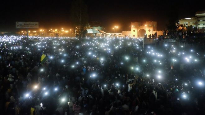 Толпа, использующая огни своих мобильных телефонов, сидит в военном штабе в Хартуме, Судан, в воскресенье, 7 апреля 2019 года