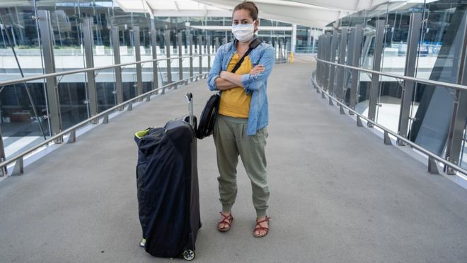 Mulher de máscara, braços cruzados e ao lado de mala em corredor de aeroporto