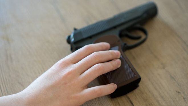Mão de criança encosta em revólver colocado sobre mesa