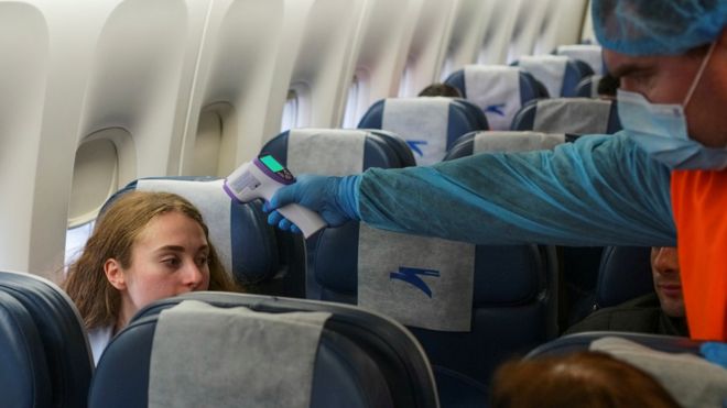 Медицинский работник в защитном снаряжении идет внутри самолета, измеряя температуру тела пассажиров, прибывших рейсом из Нью-Йорка