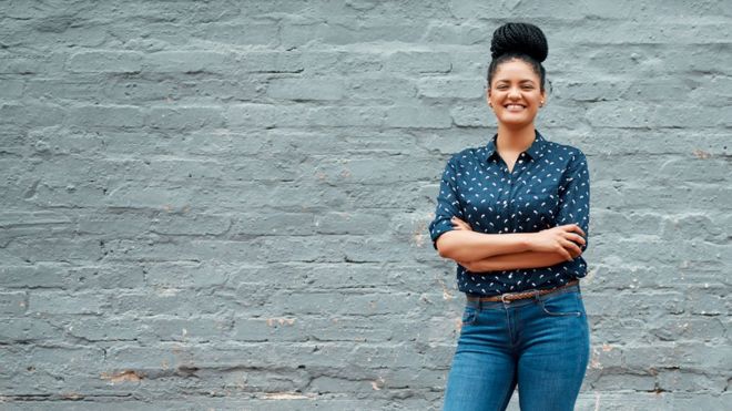 Mulher de camisa, calÃ§a-jeans, cabelos tranÃ§ado preso no alto, sorrindo, com braÃ§os cruzados e em frente a uma parede