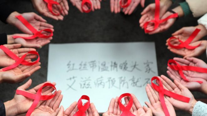 На этом снимке, сделанном 30 ноября 2015 г., изображены добровольцы с красными лентами над листом бумаги, написанным на китайском языке, на котором написано: «Красные ленты приносят тепло всем, чтобы предотвратить СПИД»