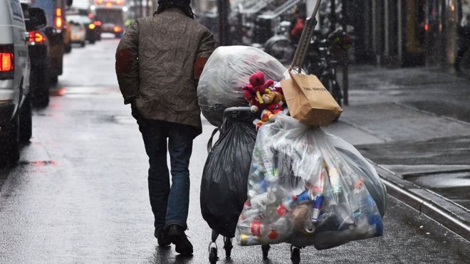 На фотографии изображен мужчина, тянущий телегу с пакетами бутылок через улицу в Нью-Йорке