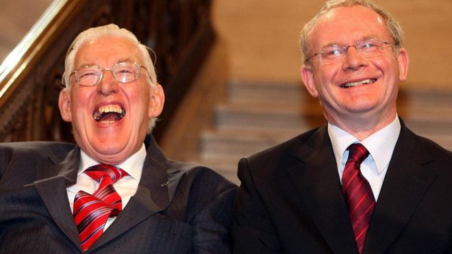 Ян Пейсли и Мартин МакГиннесс улыбаются, будучи приведенными к присяге в качестве первого и заместителей первых министров Ассамблеи Северной Ирландии