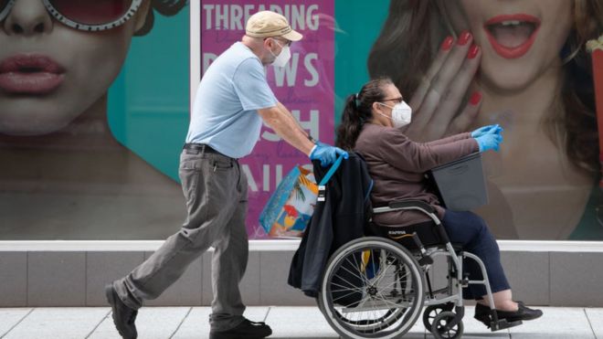 A woman sitting in a wheelchair wearing a mask, with a man in a mask pushing the chair
