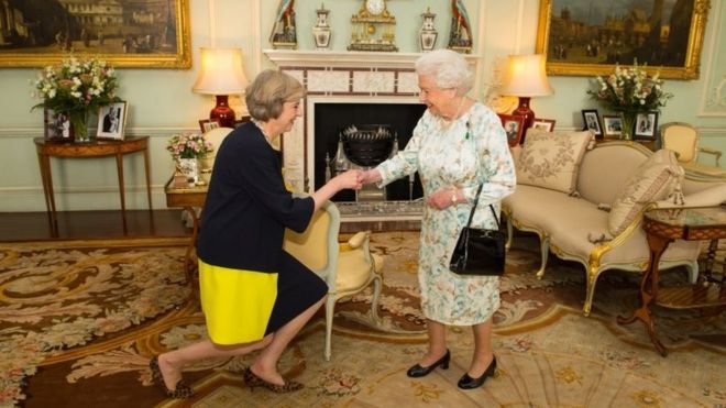 Королева Елизавета II приветствует Терезу Мэй (видно, как она кланяется, выполняет реверанс - официальное приветствие, сделанное сгибанием коленей одной ногой перед другой) в начале аудитории в Букингемском дворце в Лондоне