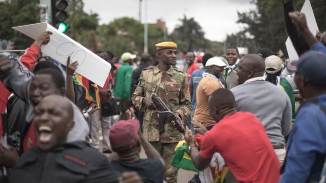 Протестующие, требующие президента Роберта Мугабе, встают, поднимают глаза и приветствуют, когда над толпой пролетает армейский вертолет, когда они собираются перед военным оцеплением на дороге, ведущей к государственному дому в Хараре, Зимбабве, суббота, 18 ноября 2017 года.
