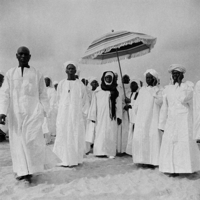 Des hommes vêtus de boubous blancs et de turbans marchent ensemble. L'un d'eux est protégé du soleil par un autre homme qui tient un parasol au-dessus de sa tête.