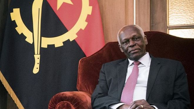 Президент душ Сантуш сидит в кресле с ангольским флагом на заднем плане