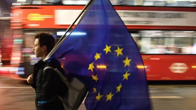 Una persona caminando con una bandera de la Unión Europea. Al fondo, un tradicional autobús de dos pisos británico pasa