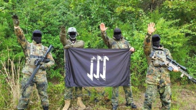 Мужчины в военной форме и масках с неонацистскими флагами в сельской местности