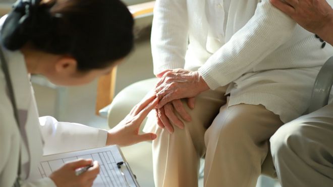Médica de costas e sentada examina joelho de idosa sentada
