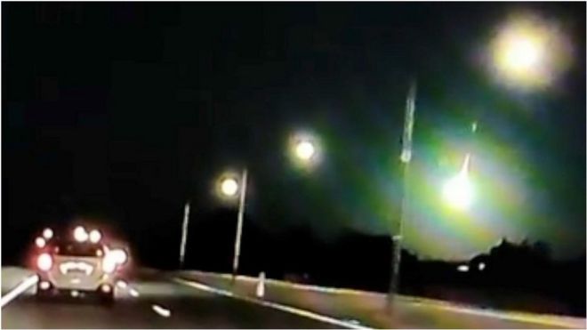 Green light flashes in dark sky above cars on road