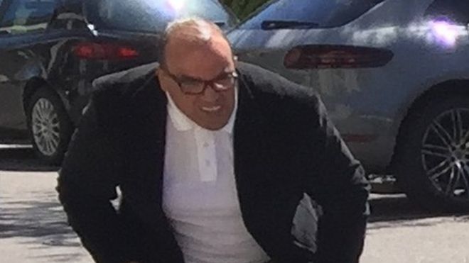 Лысеющий мужчина в очках, замеченный в белой рубашке поло под темным пиджаком, наклоняется, чтобы посмотреть на заднюю часть автомобиля