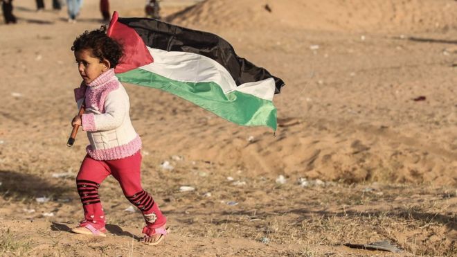 Палестинские девушки бегут с национальным флагом, когда протестующие сжигают шины на месте протеста в палатке 8 апреля 2018 года на границе Израиля и Газы к востоку от Рафаха в южной части сектора Газа