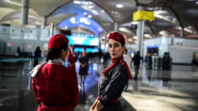 Двое сотрудников Turkish Airlines в отличительной красной униформе стоят в новых сияющих залах аэропорта, и их виднеют простирающиеся позади них. Одна спиной к камере, а другая повернулась, чтобы посмотреть прямо на фотографа