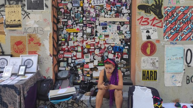 Женщина сидит перед стеной, покрытой наклейками и граффити в Сантьяго