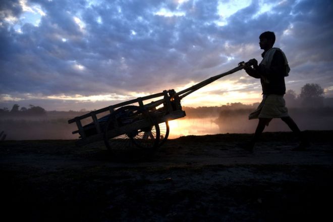 Человек толкает свою тележку, чтобы отправиться на работу рано утром во время восхода солнца, когда облака собираются на небе на окраине Гувахати в Ассаме, Индия, в пятницу, 21 декабря 2018 года