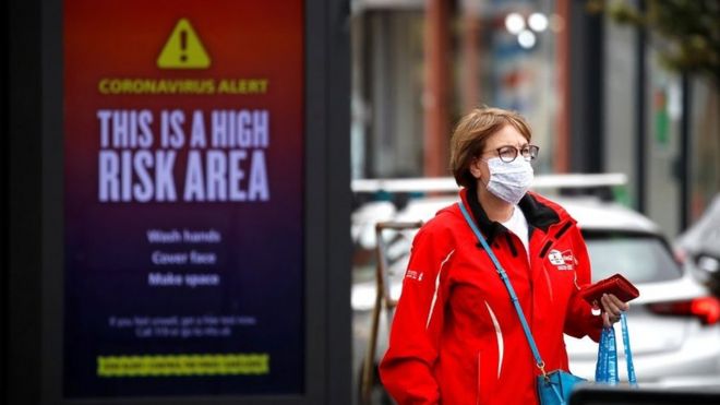 Женщина в защитной маске проходит мимо предупреждающего знака в Большом Манчестере