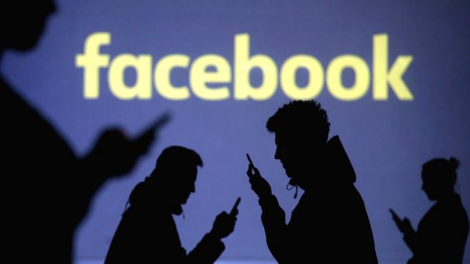 Силуэты мобильных пользователей рядом с экранной проекцией логотипа Facebook, 28 марта 2018 года