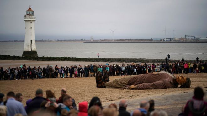 Люди собираются, чтобы посмотреть на затонувшего гиганта театральной компании Royal De Luxe, одну из уличных марионеток, принимающих участие в «Ливерпульской мечте», которые спят на пляже возле маяка Перх Рок в Нью-Брайтоне