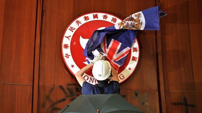 Протестующий прикрывает герб Гонконга британским колониальным флагом