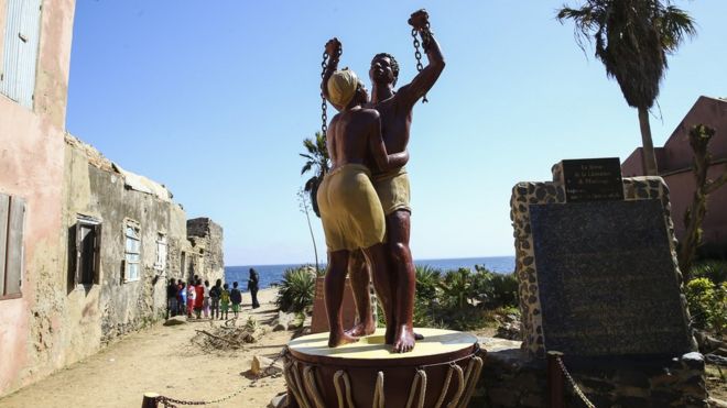 Grupo de crianças e um adulto são vistos perto da estátua de emancipação na ilha de Goree, no Senegal, que estava no coração do comércio atlântico de escravos entre os séculos XV a XIX