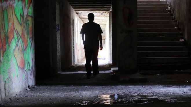 A lone figure walks down a dark hallway in the ruined building, with graffiti on the walls