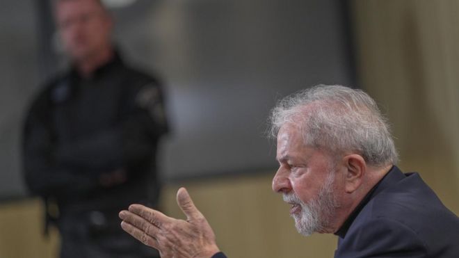 O ex-presidente Lula aparece de perfil falando, com agente de segurança no plano de fundo