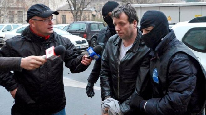 Марселя Лазаря Лехеля сопровождают полицейские в масках в Бухаресте после ареста 22 января 2014 года.