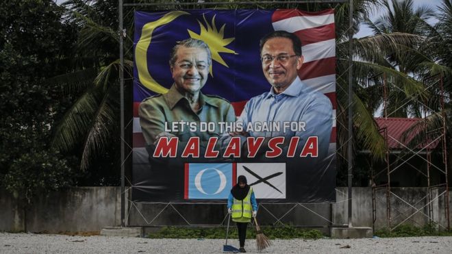 Перед рекламным щитом виден рабочий, на котором изображены бывший премьер-министр Малайзии Махатхир Мохамад, а также бывший заместитель премьер-министра и бывший лидер оппозиционной партии Анвар Ибрагим