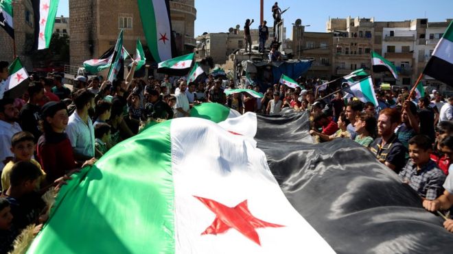 Сирийцы держат государственный флаг оппозиции на акции протеста в удерживаемом повстанцами городе Идлиб (28 сентября 2018 года)