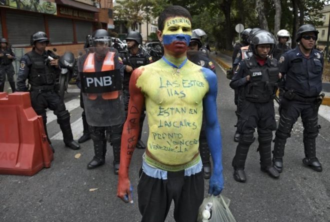 Человек с телом, выкрашенным в венесуэльский национальный флаг " чтение цветов "Звезды на улицах. Мы скоро будем победоносной Венесуэлой », которая демонстрируется перед полицией по охране общественного порядка во время демонстрации оппозиции около авиабазы ??Ла-Карлота в Каракасе 4 мая 2019 года