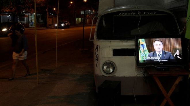 Pronunciamento de Bolsonaro é exibido em TV no meio da rua à noite no Rio de Janeiro, ao lado de combe e pedestre