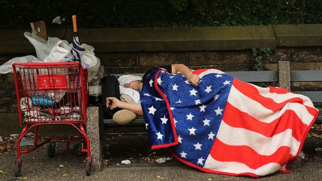 Un hombre durmiendo en un banco con una manta con el dibujo de la bandera de Estados Unidos